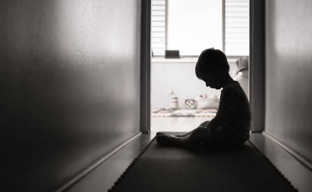 اندھیرے کمرے میں موجود ایک کم عمر بچہ دیوار کے قریب اداس بیٹھے ہوئے