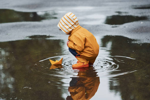 بارش کے پانی میں کاغذ کی کشتی سے کھیلتا ہوا چھوٹا بچہ