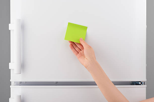 Female hand holding blank green sticky paper note on white refrigerator door