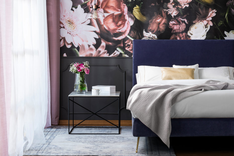 Flowers on table next to pink bed with grey sheets in bedroom interior with wallpaper.