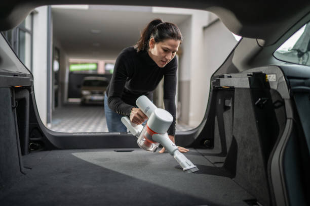 Young woman standing and cleaning car trunk with hand vacuum cleaner